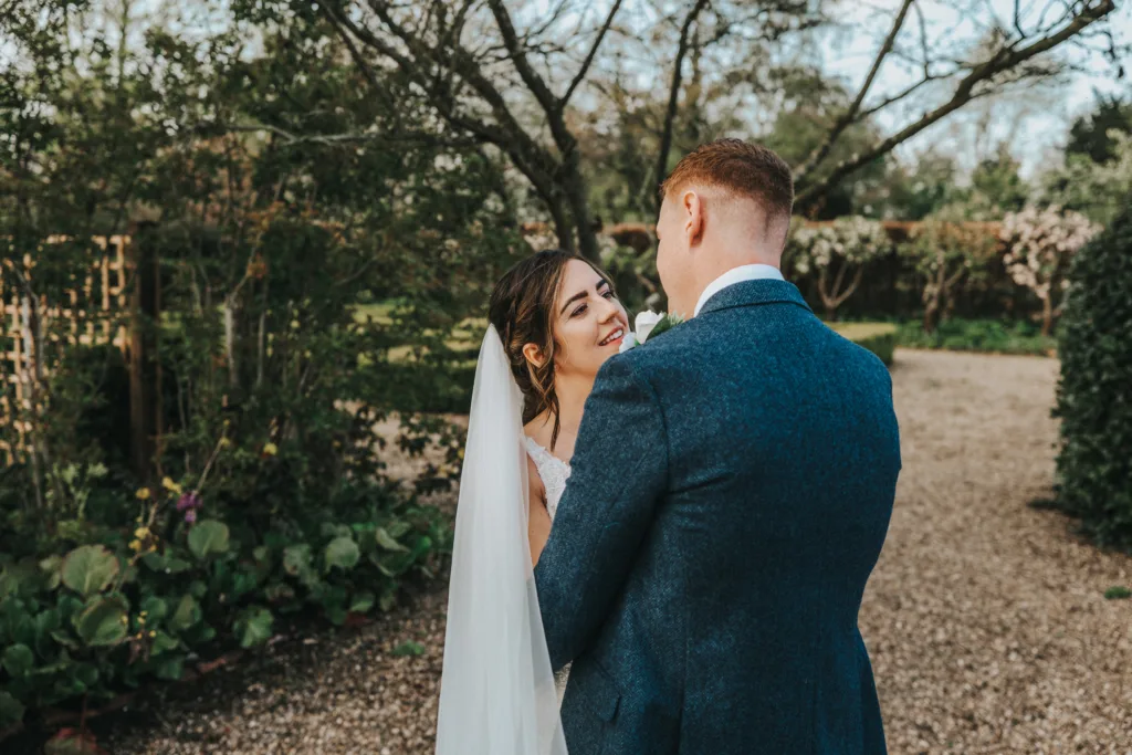 A bride and groom stand outdoors facing each other, smiling lovingly. The bride wears a white dress and veil; the groom is in a blue suit. They are surrounded by greenery and trees on a gravel path. © Aimee Lince Photography, wedding and lifestyle photographer in Lincolnshire