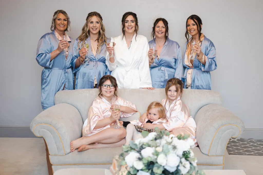 Seven women and girls in matching dressing gowns smile and hold drinks while posing together in a living room. Three children sit on a beige sofa; four adults stand behind them. A flower arrangement is visible in the foreground. © Aimee Lince Photography, wedding and lifestyle photographer in Lincolnshire