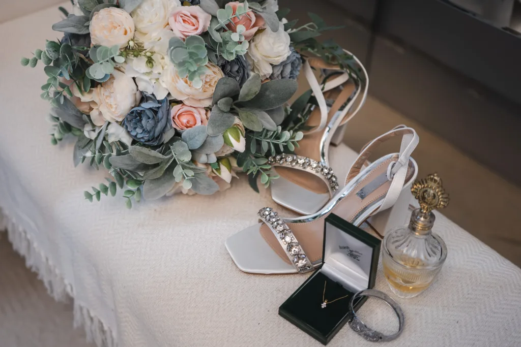 A bridal bouquet, jewelled white high-heeled shoes, a gold necklace in a green box, a sparkling bracelet, and a vintage-style perfume bottle arranged on a light fabric surface. © Aimee Lince Photography, wedding and lifestyle photographer in Lincolnshire