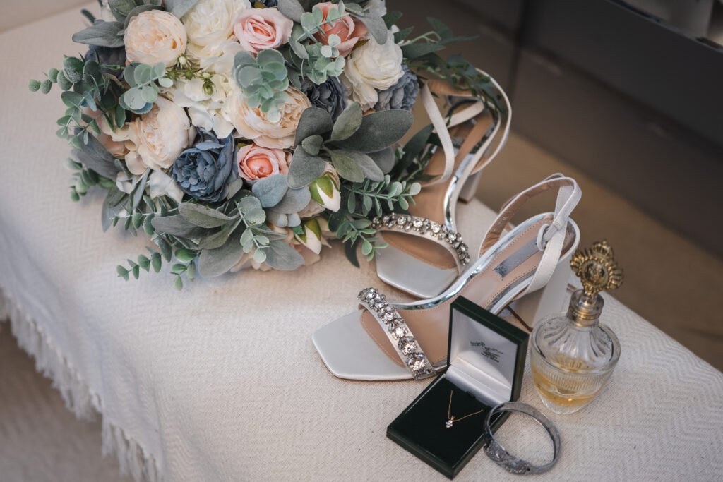 A bridal bouquet, jewelled white high-heeled shoes, a gold necklace in a green box, a sparkling bracelet, and a vintage-style perfume bottle arranged on a light fabric surface. © Aimee Lince Photography, wedding and lifestyle photographer in Lincolnshire