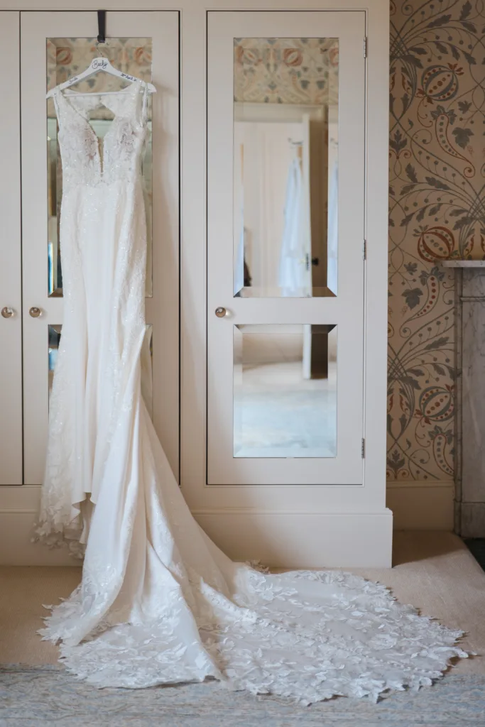 A white lace wedding dress with a long train hangs from a personalised hanger on a mirrored wardrobe in a softly lit room with patterned wallpaper. © Aimee Lince Photography, wedding and lifestyle photographer in Lincolnshire