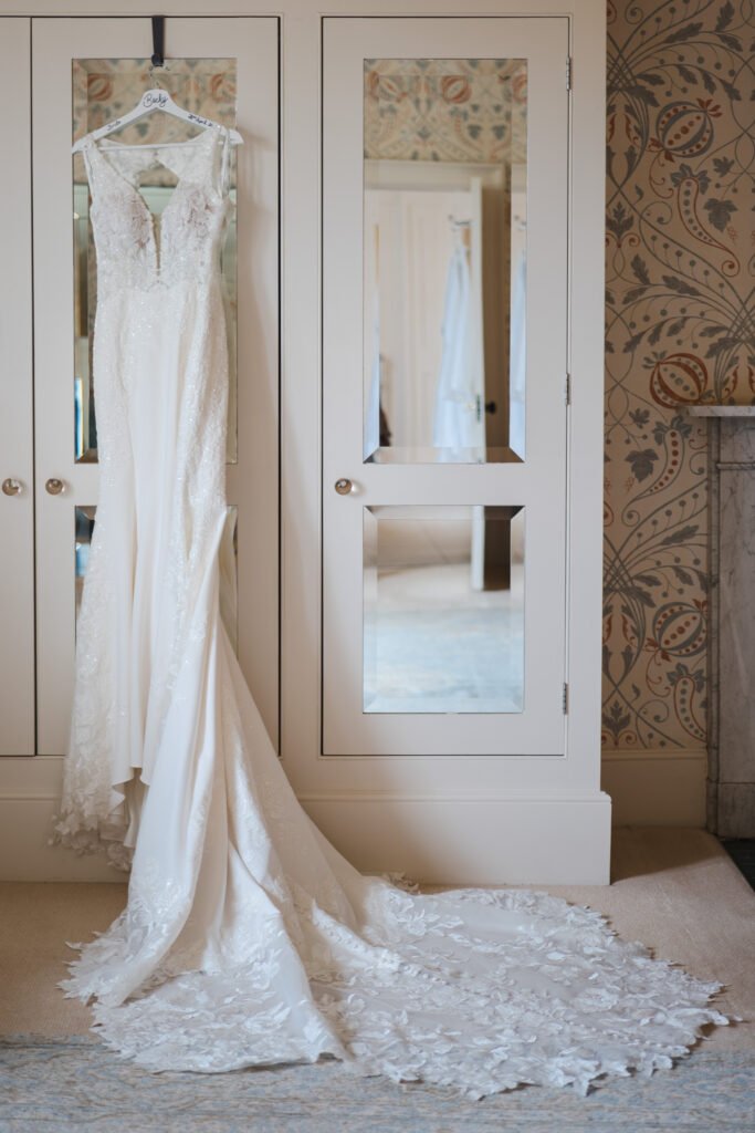 A white lace wedding dress with a long train hangs from a personalised hanger on a mirrored wardrobe in a softly lit room with patterned wallpaper. © Aimee Lince Photography, wedding and lifestyle photographer in Lincolnshire