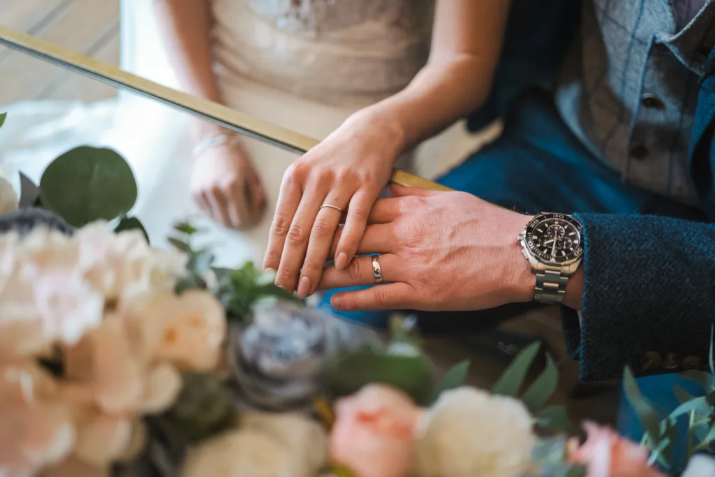 A close-up of a couple’s hands resting on a table, both wearing wedding rings. The scene includes a bouquet of flowers and the man is wearing a wristwatch. © Aimee Lince Photography, wedding and lifestyle photographer in Lincolnshire