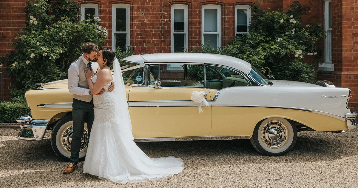 A bride in a white dress and veil embraces a groom in a waistcoat and tie. Captured by a wedding photographer, they stand beside a vintage yellow and white car, in front of a brick building with white-trimmed windows and green shrubbery. © Aimee Lince Photography, Lincolnshire photographer