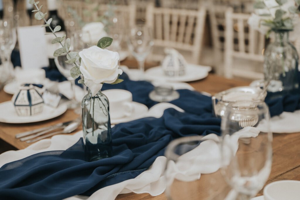 A wooden table is elegantly set with white crockery, silver cutlery, and clear glassware. A navy blue and white fabric runner decorates the table, featuring glass vases with white roses and greenery as centrepieces. White chairs are arranged around the table. © Aimee Lince Photography, Lincolnshire photographer