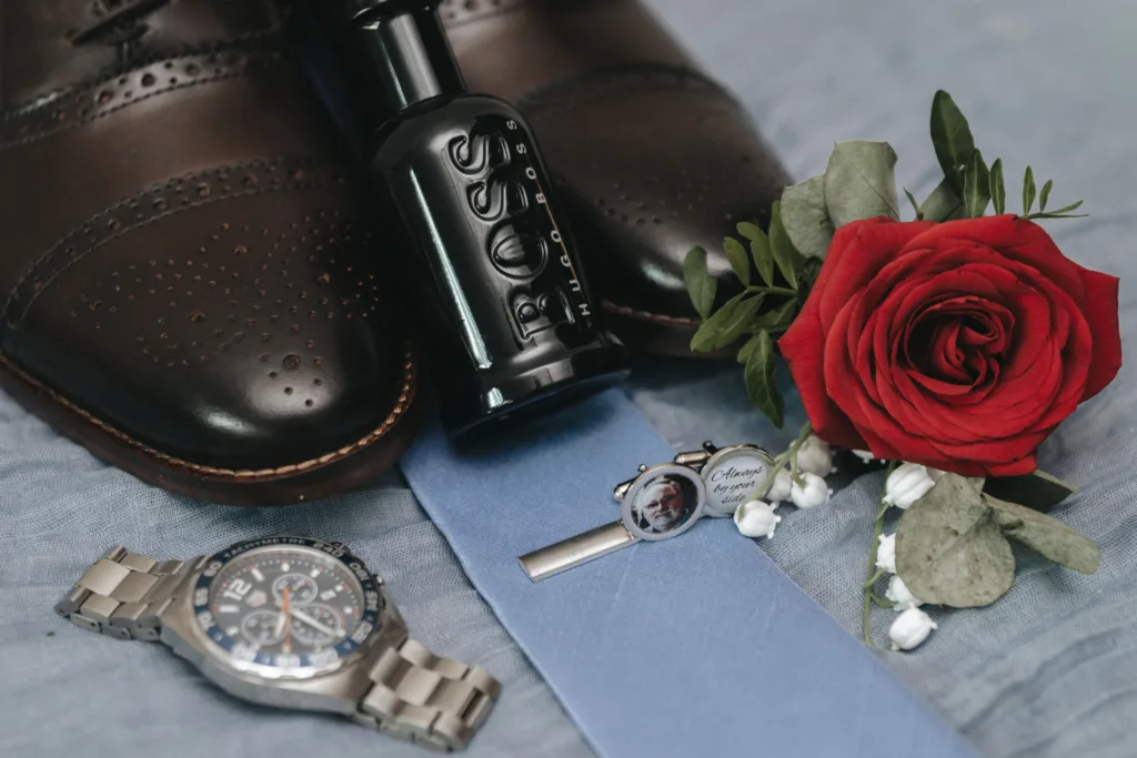 A close-up of wedding accessories: brown leather shoes, a BOSS aftershave bottle, a silver wristwatch, a blue tie with a tie clip, and a buttonhole with a red rose and greenery, all arranged on a light fabric. © Aimee Lince Photography, wedding and lifestyle photographer in Lincolnshire
