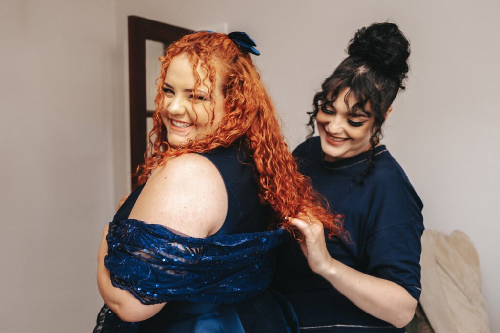 Two women smile as one helps the other adjust a sparkly blue shawl. The woman in front has long curly red hair and wears a blue dress, while the other has dark curly hair in a bun and wears a navy top. They appear happy, standing indoors. © Aimee Lince Photography, Lincolnshire photographer