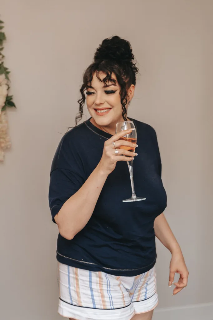 A smiling woman with curly dark hair in a bun holds a glass of rosé wine. She wears a navy blue T-shirt and striped shorts in pastel colours, standing against a light, plain background with a touch of floral decor on the side. © Aimee Lince Photography, Lincolnshire photographer
