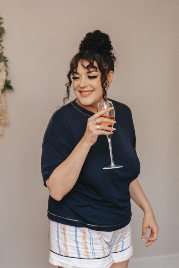 A smiling woman with curly dark hair in a bun holds a glass of rosé wine. She wears a navy blue T-shirt and striped shorts in pastel colours, standing against a light, plain background with a touch of floral decor on the side. © Aimee Lince Photography, Lincolnshire photographer