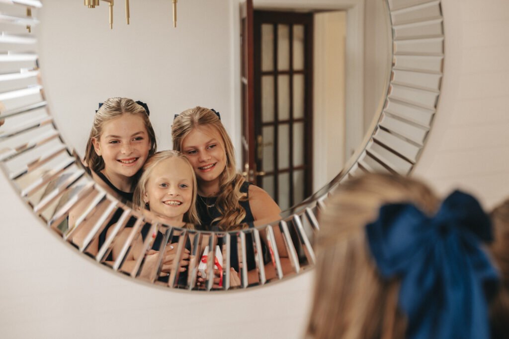 Three smiling girls with blonde hair, wearing dark outfits, are reflected in a round, decorative mirror. One girl in the foreground has a large blue bow in her hair. They appear happy and close together in a warmly lit room. © Aimee Lince Photography, Lincolnshire photographer