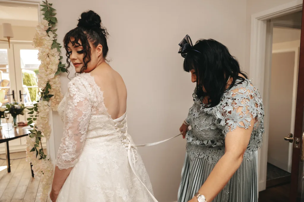 A bride in a lace wedding dress stands as another woman in a blue lace dress ties the back of her gown. Both have dark hair; the bride looks over her shoulder, and the room is softly lit, decorated with white flowers and natural light. © Aimee Lince Photography, Lincolnshire photographer