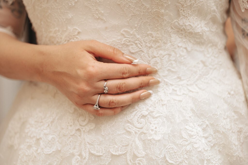 A close-up of a hand with a French manicure gently resting on a white lace wedding dress. The hand wears an engagement ring and a wedding band, highlighting intricate lace patterns on the dress. © Aimee Lince Photography, Lincolnshire photographer