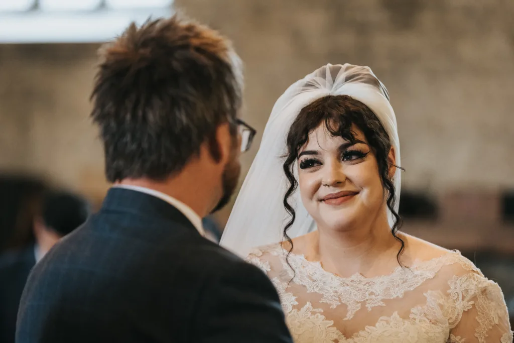 A bride in a lace wedding dress and veil smiles warmly at a man in glasses and a suit, who is seen from behind. They are indoors, possibly at a wedding ceremony, with soft, natural light and blurred background. © Aimee Lince Photography, Lincolnshire photographer