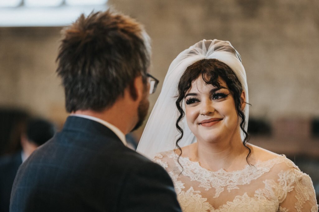 A bride in a lace wedding dress and veil smiles warmly at a man in glasses and a suit, who is seen from behind. They are indoors, possibly at a wedding ceremony, with soft, natural light and blurred background. © Aimee Lince Photography, Lincolnshire photographer