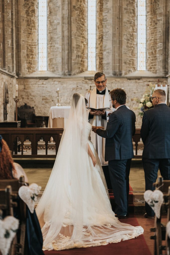 A bride in a long white dress and veil stands facing a groom in a suit before a vicar in a stone church with stained glass windows. The vicar holds a book, smiles, and presides over the wedding ceremony as seated guests look on. © Aimee Lince Photography, Lincolnshire photographer