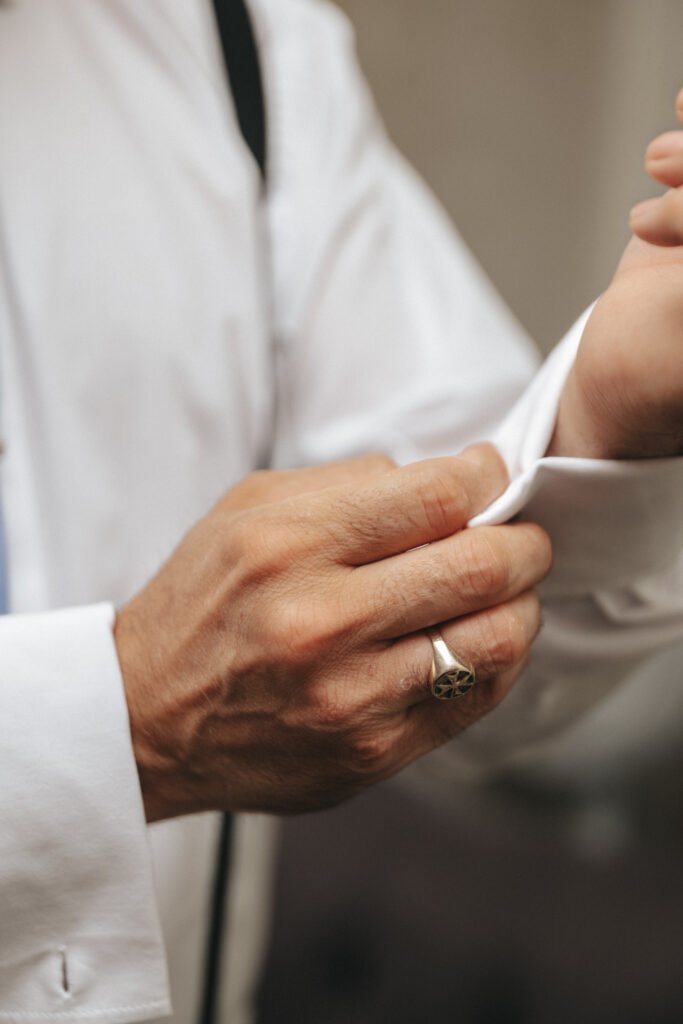 A close-up of a person fastening the cuff of a white dress shirt. The person is wearing a ring with a geometric design and black braces, and the background is blurred. © Aimee Lince Photography, wedding and lifestyle photographer in Lincolnshire