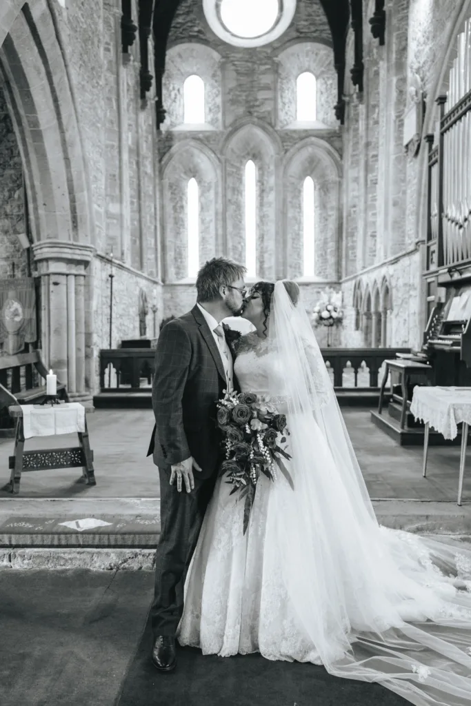 A bride and groom kiss in a historic stone church. The bride wears a long veil and lace gown, holding a bouquet, while the groom is in a suit. Light streams through tall arched windows behind them. The photo is in black and white. © Aimee Lince Photography, Lincolnshire photographer