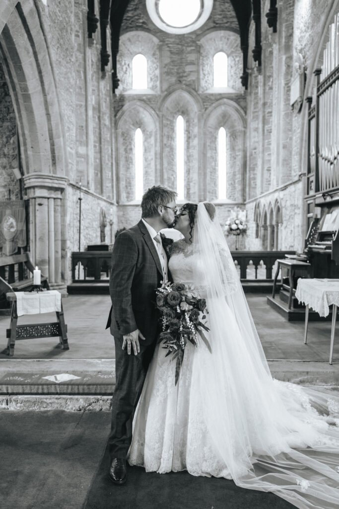 A bride and groom kiss in a historic stone church. The bride wears a long veil and lace gown, holding a bouquet, while the groom is in a suit. Light streams through tall arched windows behind them. The photo is in black and white. © Aimee Lince Photography, Lincolnshire photographer