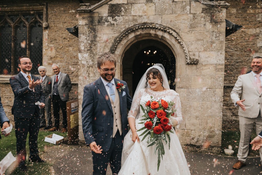 A bride in a white lace gown and a groom in a blue suit walk outside a stone church, smiling, whilst guests throw confetti. The bride holds a bouquet of red roses. Several guests in suits stand nearby, some mid-gesture, celebrating the couple. © Aimee Lince Photography, Lincolnshire photographer