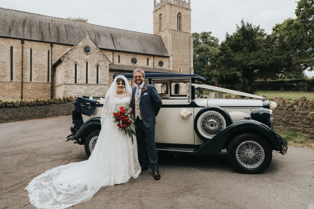A bride in a white gown and veil holding a red bouquet stands beside a groom in a blue suit, both smiling in front of a vintage cream and navy car. Behind them is St. Peter's Church in Bottesford, a stone church with a tall bell tower and trees in the background. © Aimee Lince Photography, Lincolnshire photographer