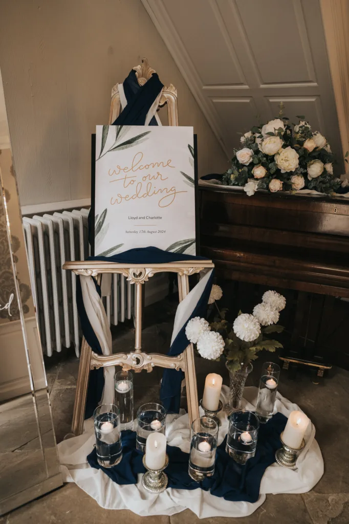 An easel displays a "Welcome to Our Wedding" sign draped in navy and white fabric. Below, glass vases with floating candles, ivory flowers, and votive candles are arranged on the floor. A floral arrangement sits on a piano in the background. © Aimee Lince Photography, Lincolnshire photographer