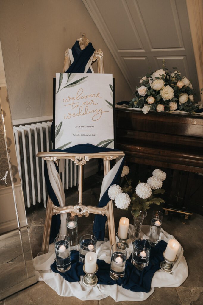 An easel displays a "Welcome to Our Wedding" sign draped in navy and white fabric. Below, glass vases with floating candles, ivory flowers, and votive candles are arranged on the floor. A floral arrangement sits on a piano in the background. © Aimee Lince Photography, Lincolnshire photographer