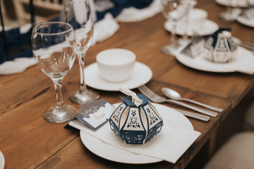 A close-up of a wooden table laid for a meal, featuring white plates, silver cutlery, wine glasses, a small white bowl, and a blue and white decorative lantern-shaped party favour on a serviette, with a place card and neatly folded blue cloth in the background. © Aimee Lince Photography, Lincolnshire photographer