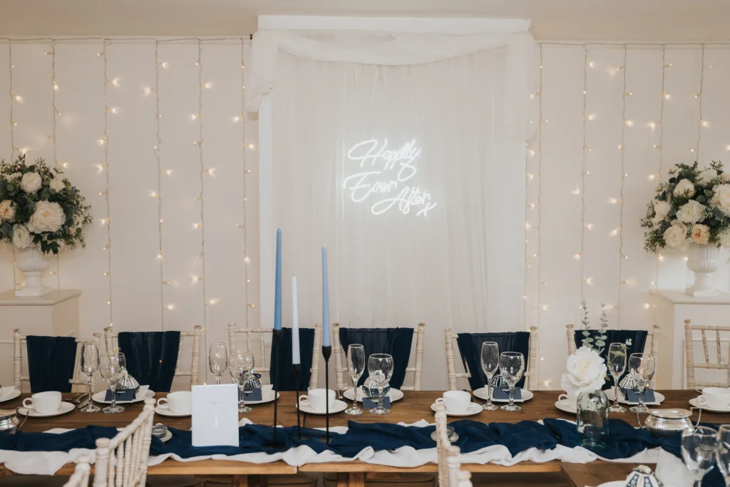 A wedding reception table is elegantly set with navy blue and white decor, tall blue candles, white flowers in vases, glassware, and place settings. Behind the table, fairy lights and white drapery with a neon sign reading “Happily Ever After” are displayed. © Aimee Lince Photography, Lincolnshire photographer