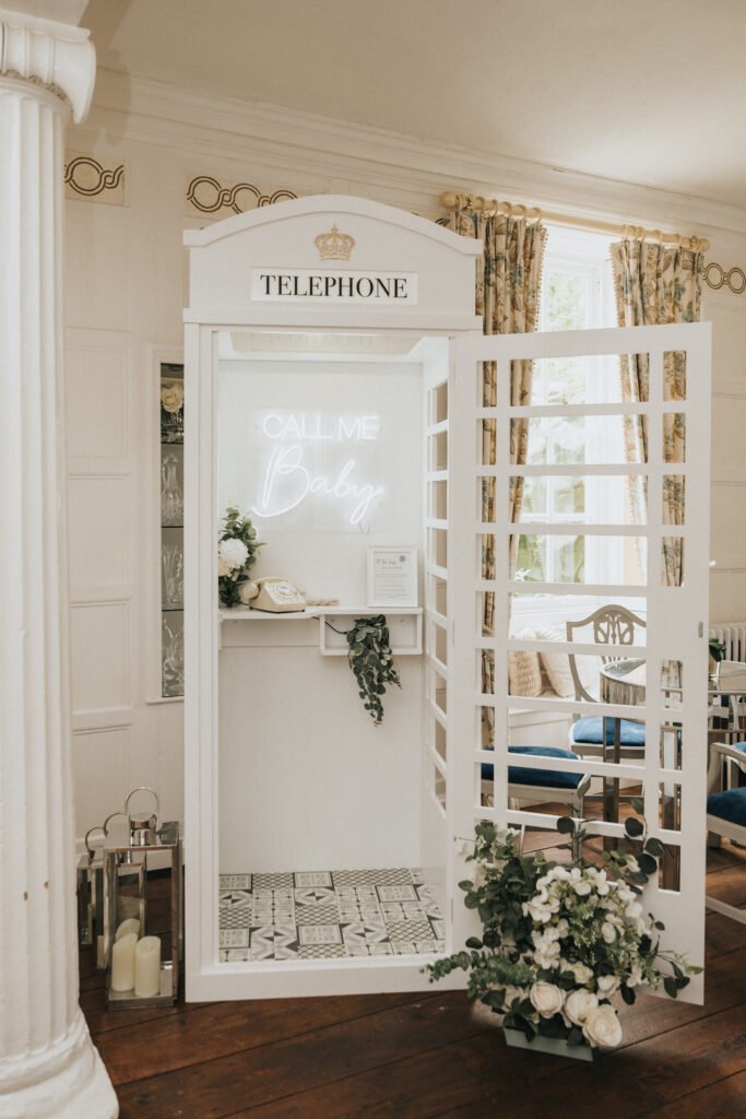 White vintage-style phone box with “TELEPHONE” sign. Inside, a neon sign reads “Call Me Baby.” The box is decorated with greenery, flowers, and a guest book, with candles and more flowers beside it. Elegant room with classic decor and patterned floor tiles. © Aimee Lince Photography, Lincolnshire photographer
