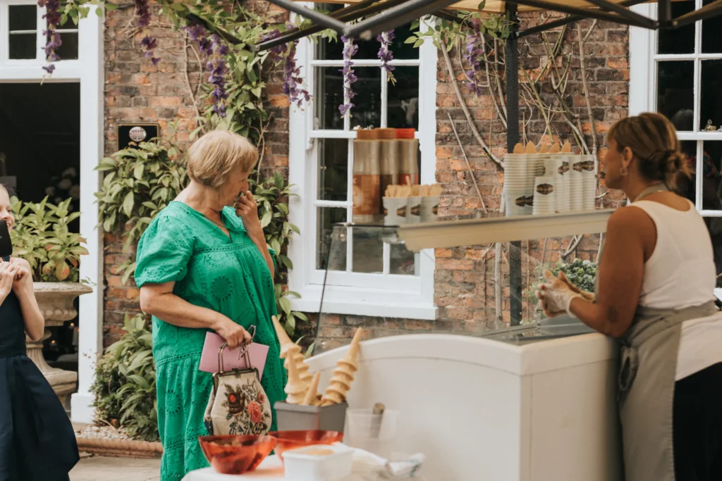 A woman in a green dress stands in front of an outdoor ice cream stall, holding a pink handbag and floral bag. She looks at the vendor, who is behind the counter. Swirled cones, tubs, and plants decorate the scene, with brick and white windows in the background. © Aimee Lince Photography, Lincolnshire photographer