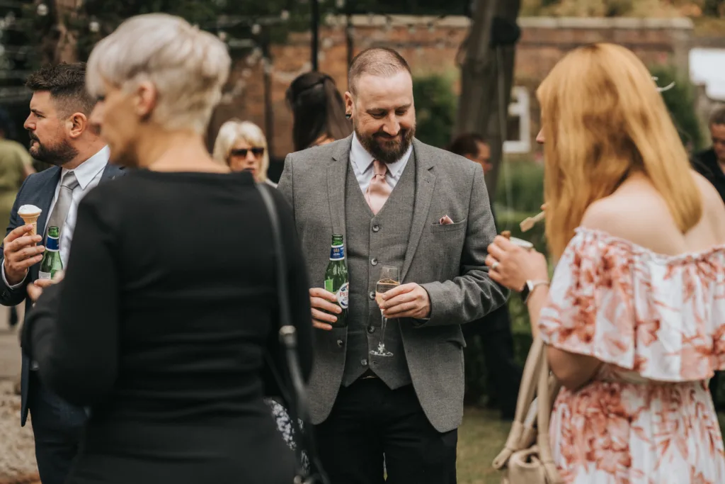 A group of people dressed in formal attire stand outdoors at a social gathering. A bearded man in a grey suit holds a beer and a wine glass, smiling, while others nearby talk and hold drinks. The background shows a brick wall and greenery. © Aimee Lince Photography, Lincolnshire photographer