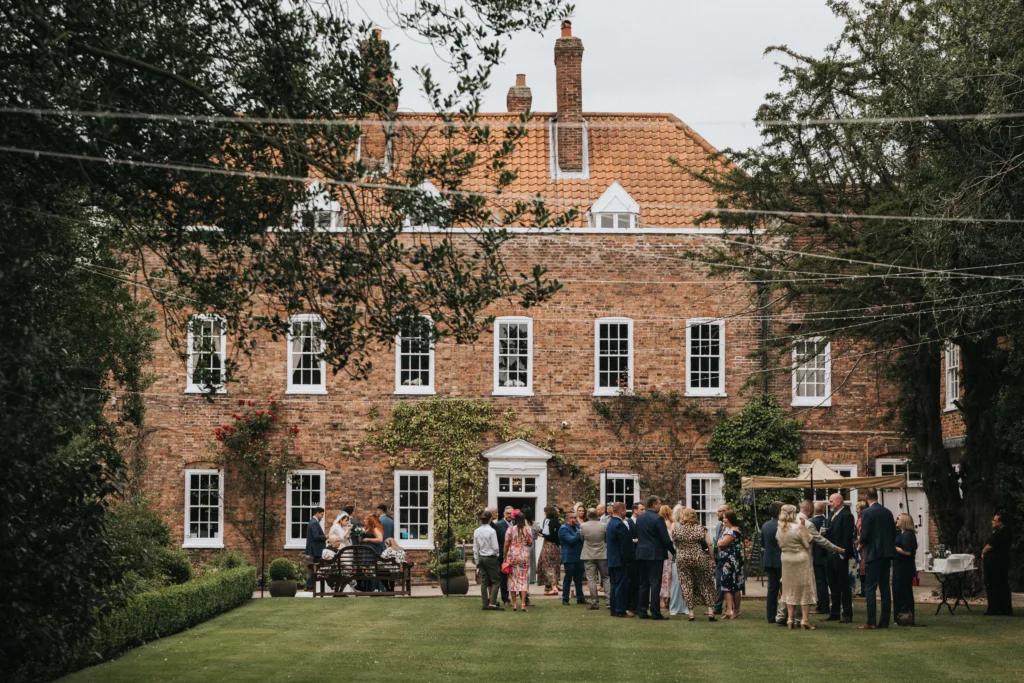 A group of people in formal attire gather and socialise on a lawn in front of a large brick house with white window frames and a red-tiled roof, surrounded by trees and greenery. © Aimee Lince Photography, wedding and lifestyle photographer in Lincolnshire