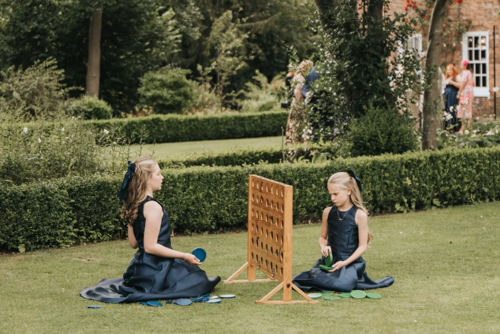 Two young girls in matching navy blue dresses sit on grass, playing a large outdoor Connect Four game. One girl looks at the game, whilst the other holds discs. Lush greenery, hedges, and a brick building with people in the background complete the scene. © Aimee Lince Photography, Lincolnshire photographer