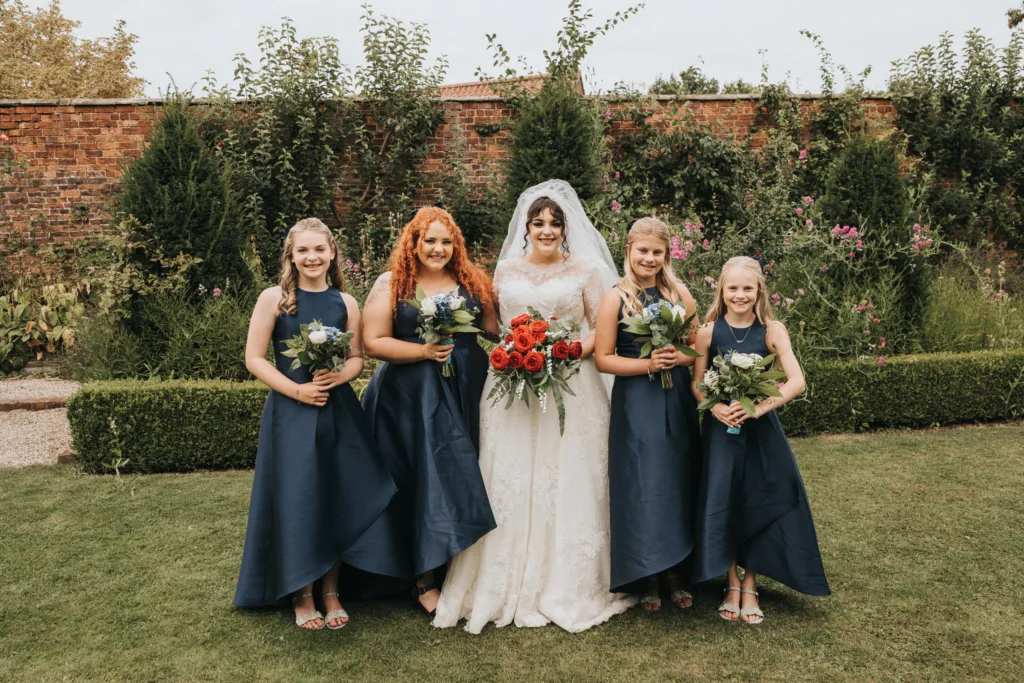 A bride in a white dress holds a red bouquet, standing with four bridesmaids in navy blue dresses, each holding green and white bouquets. They are outside on grass, in front of a brick wall and lush greenery, all smiling at the camera. © Aimee Lince Photography, Lincolnshire photographer