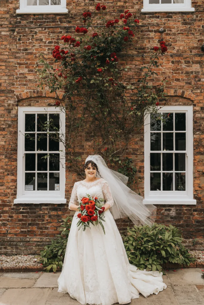 A bride in a white lace dress and veil holds a bouquet of red roses, standing in front of a brick building with white-trimmed windows and red roses climbing the wall behind her. She is smiling, and greenery frames the lower part of the scene. © Aimee Lince Photography, Lincolnshire photographer