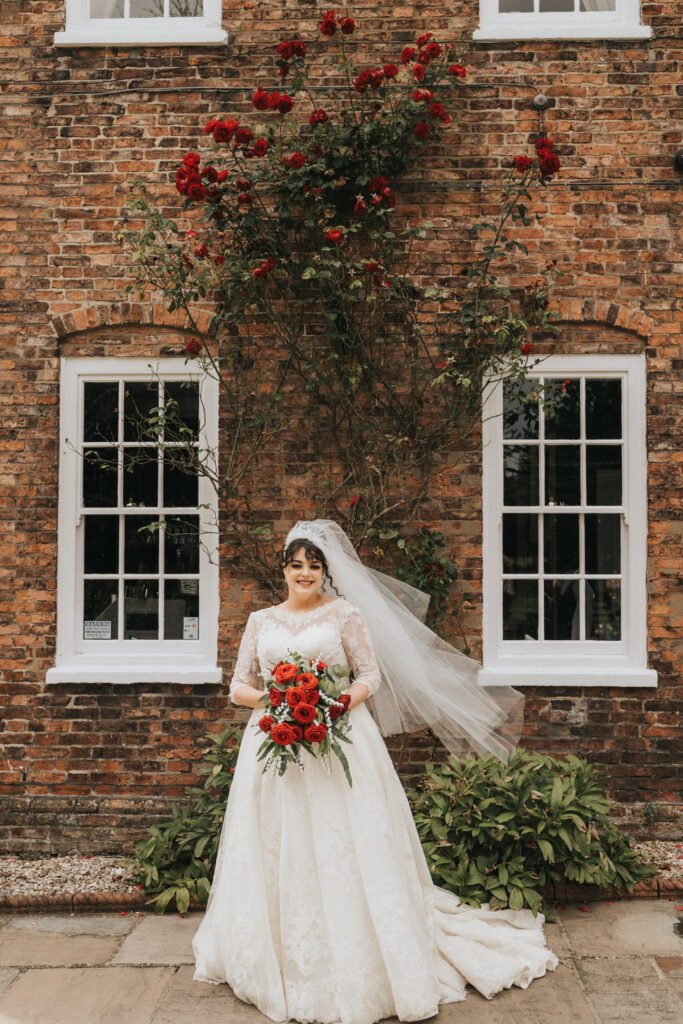 A bride in a white lace dress and veil holds a bouquet of red roses, standing in front of a brick building with white-trimmed windows and red roses climbing the wall behind her. She is smiling, and greenery frames the lower part of the scene. © Aimee Lince Photography, Lincolnshire photographer