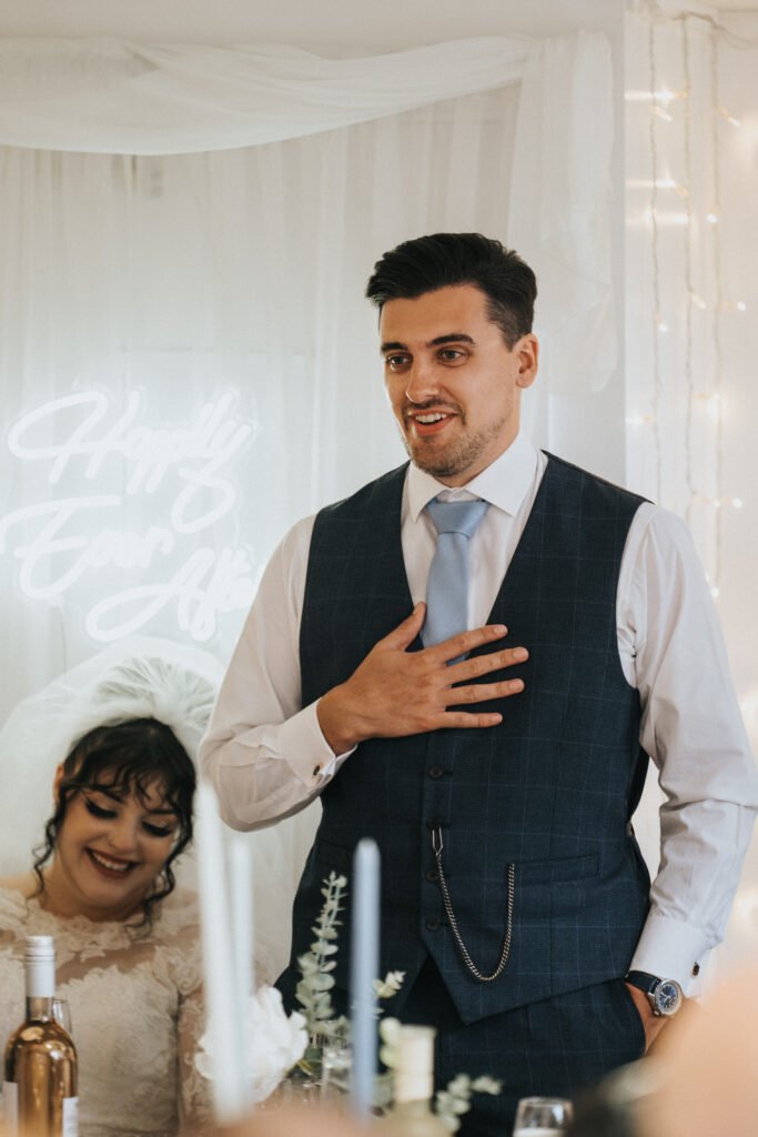 A man in a blue checked waistcoat and light blue tie stands giving a speech, hand on chest, smiling. Behind him, a woman in a wedding dress sits smiling. The background features white drapery, fairy lights, and a neon sign reading "Happily Ever After. © Aimee Lince Photography, Lincolnshire photographer