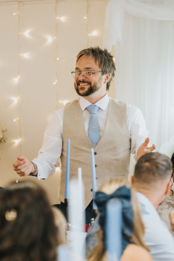 A man with glasses, a beard, and short brown hair stands and gestures while smiling. He wears a light beige waistcoat, white shirt, and light blue tie. People seated in front of him listen. Warm fairy lights and white drapes decorate the background. © Aimee Lince Photography, Lincolnshire photographer