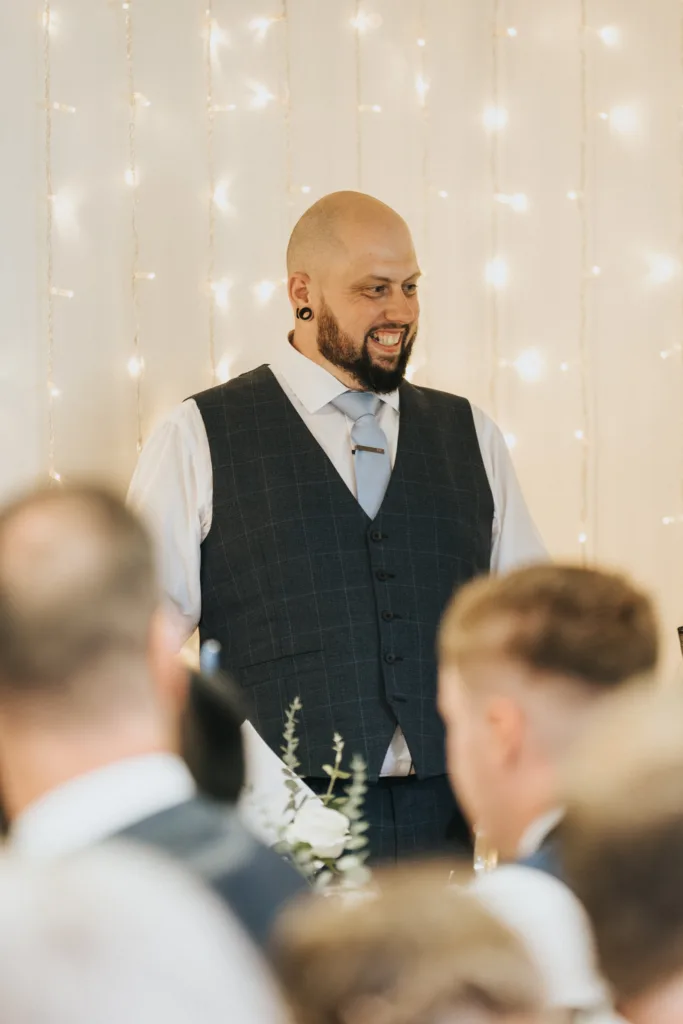 A bald man with a beard, wearing a dark checked waistcoat, white shirt, and light tie, stands smiling amid blurred guests. Behind him, warm fairy lights hang against a light-coloured wall, creating a festive, celebratory atmosphere. © Aimee Lince Photography, Lincolnshire photographer
