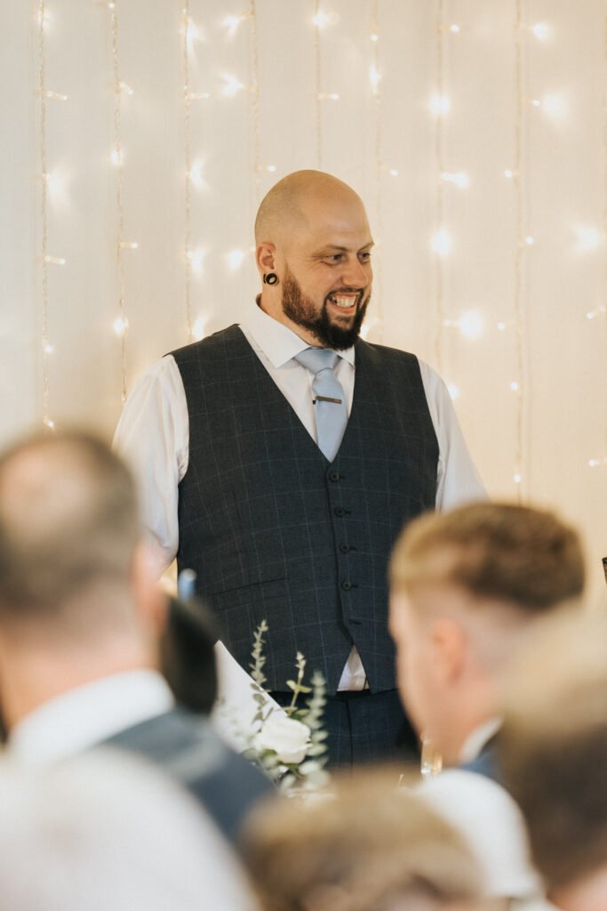 A bald man with a beard, wearing a dark checked waistcoat, white shirt, and light tie, stands smiling amid blurred guests. Behind him, warm fairy lights hang against a light-coloured wall, creating a festive, celebratory atmosphere. © Aimee Lince Photography, Lincolnshire photographer