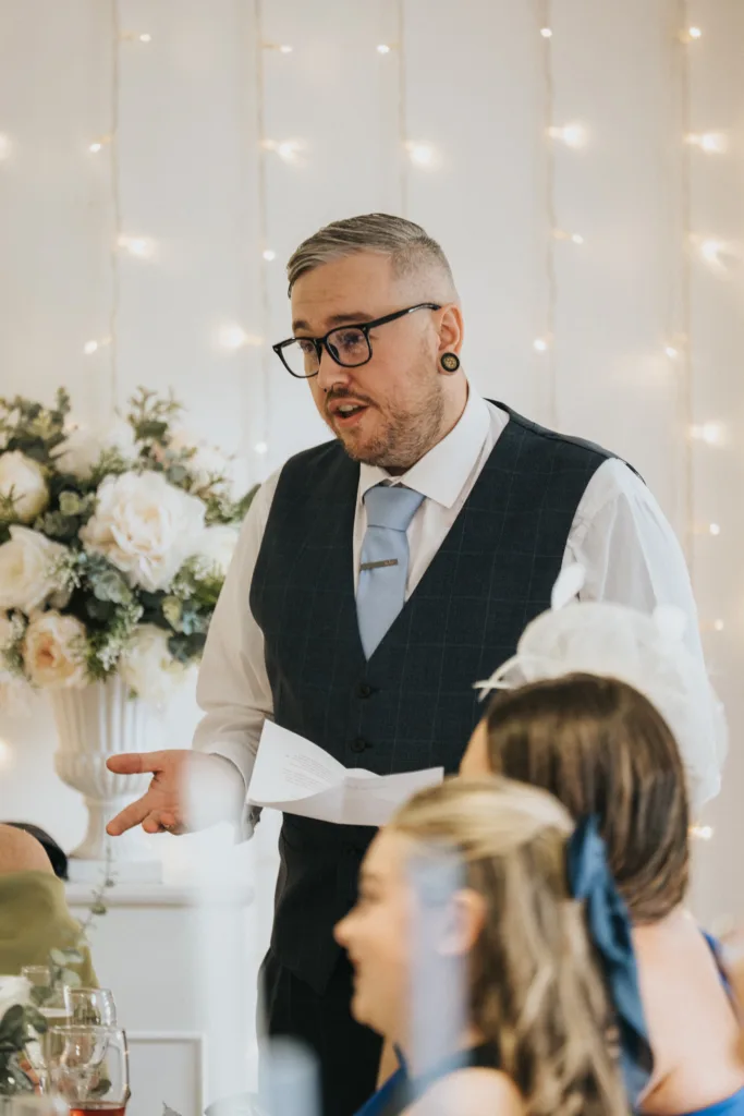 A man with glasses, short hair, and gauged ears, wearing a white shirt, light blue tie, and dark waistcoat, gives a speech whilst holding a piece of paper. He stands near women in blue dresses, with a bouquet of white flowers and fairy lights in the background. © Aimee Lince Photography, Lincolnshire photographer