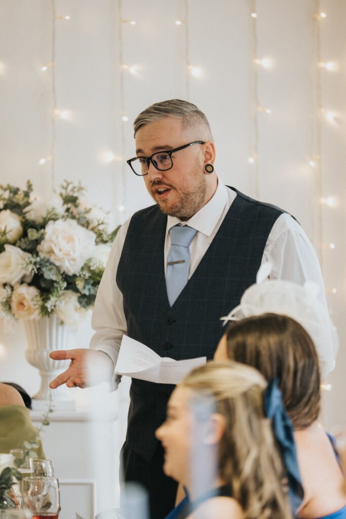 A man with glasses, short hair, and gauged ears, wearing a white shirt, light blue tie, and dark waistcoat, gives a speech whilst holding a piece of paper. He stands near women in blue dresses, with a bouquet of white flowers and fairy lights in the background. © Aimee Lince Photography, Lincolnshire photographer