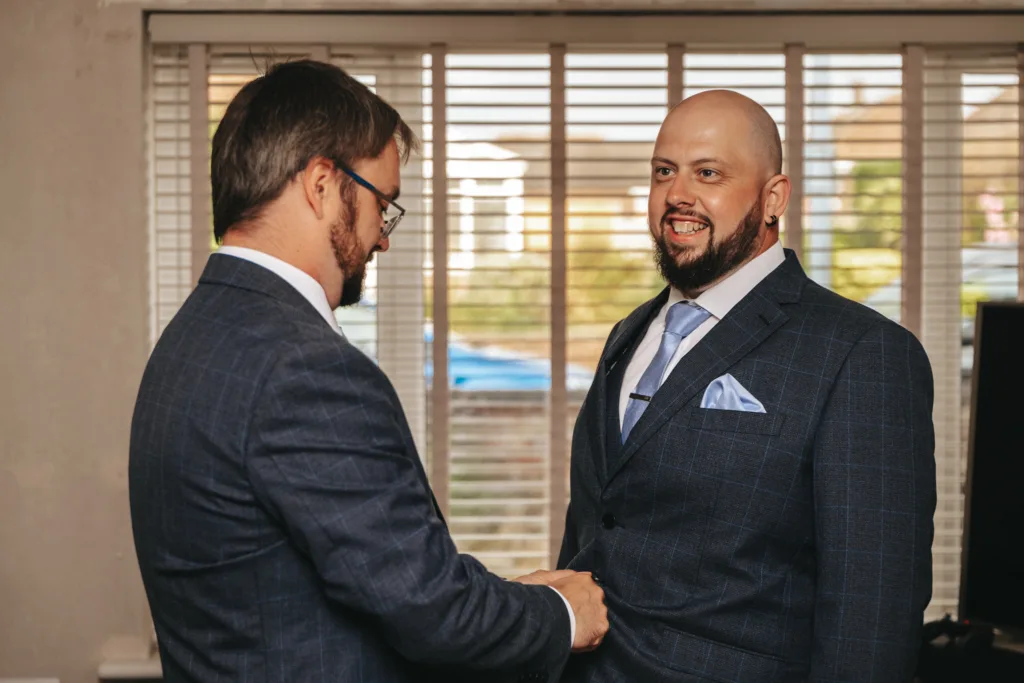 Two men in suits stand indoors by a window with blinds. One man adjusts the other’s suit jacket while they both smile, suggesting a friendly or celebratory moment. © Aimee Lince Photography, wedding and lifestyle photographer in Lincolnshire
