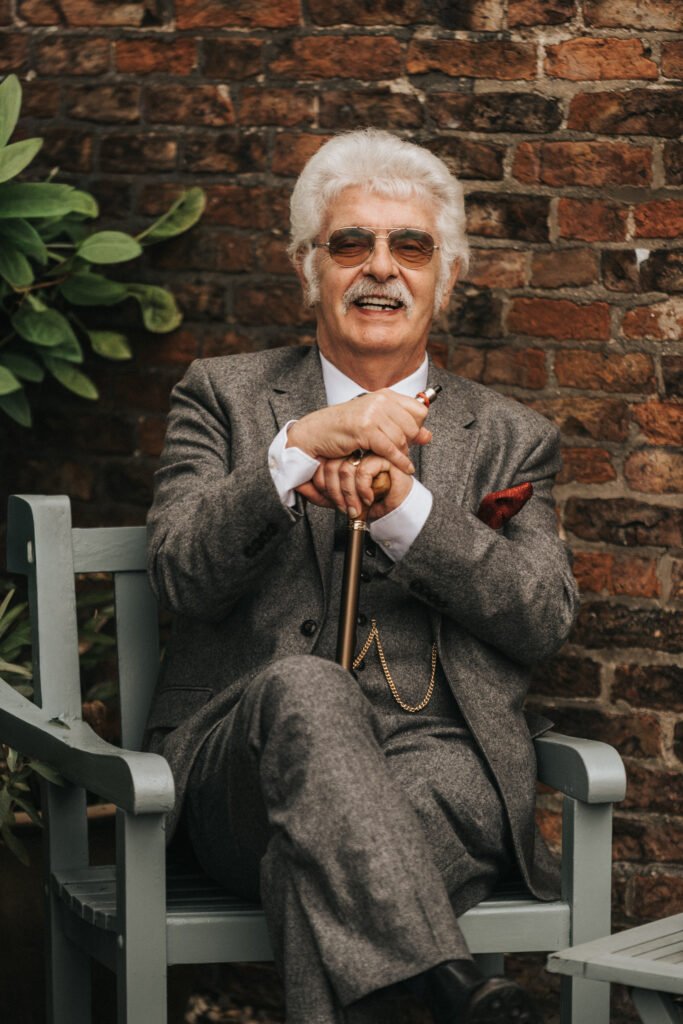 An older man with white hair and a moustache, wearing a grey suit, sunglasses, and pocket watch, sits on a bench against a brick wall. He smiles, holding a walking stick, with greenery to the side. © Aimee Lince Photography, Lincolnshire photographer