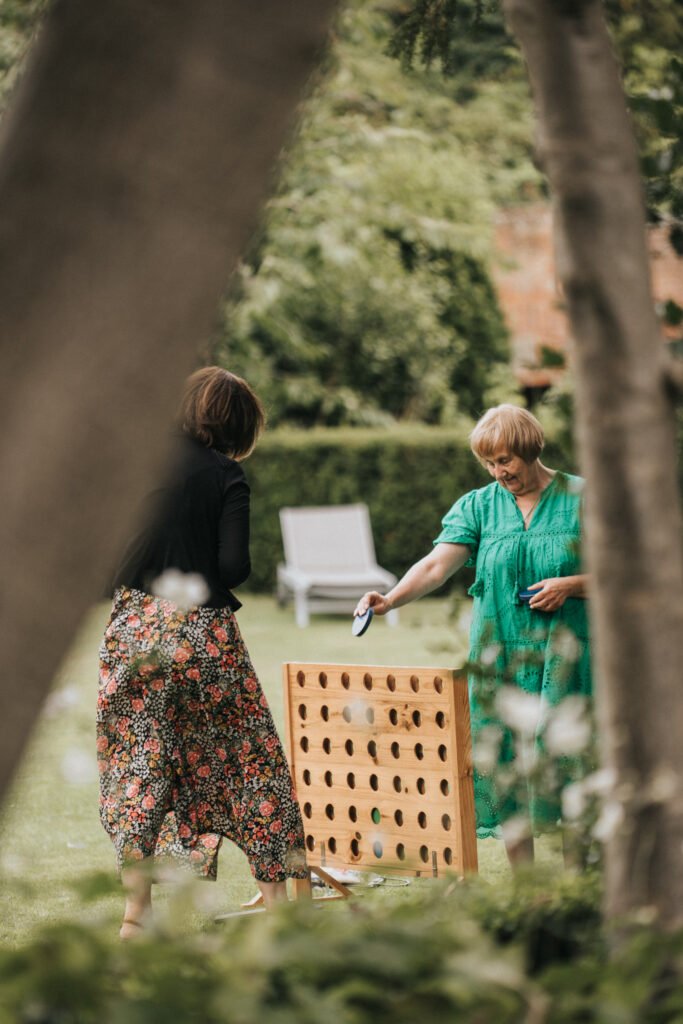 Two women stand outdoors playing a large, wooden Connect Four game on green grass. One wears a green dress, the other a black top and floral skirt. A lounger and trimmed hedges are visible in the blurred background, framed by tree trunks. © Aimee Lince Photography, Lincolnshire photographer
