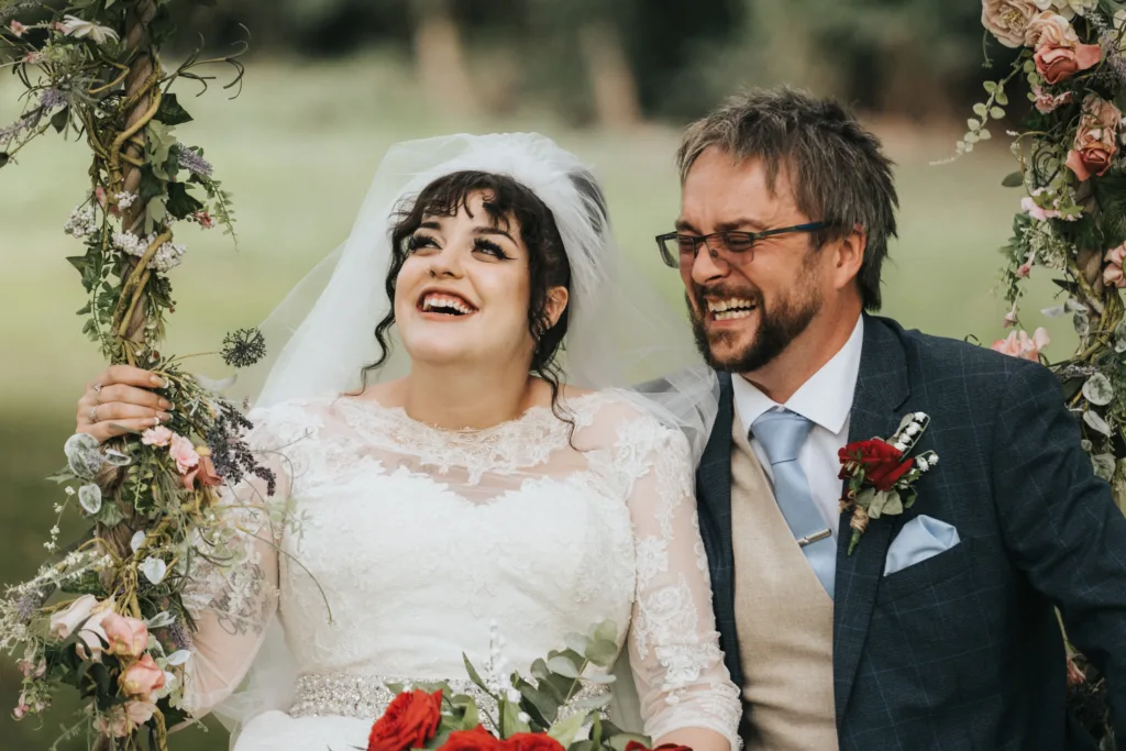 A joyful bride in a white lace dress and veil sits next to a smiling groom in a blue suit with a red buttonhole. They are surrounded by flowers and greenery, laughing together outdoors on a wedding day. © Aimee Lince Photography, Lincolnshire photographer