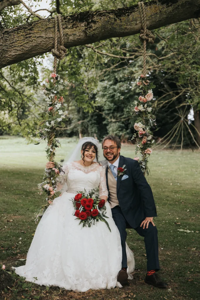 A bride in a white lace gown holding red roses and a groom in a navy suit sit together on a flower-adorned swing hanging from a tree. They are smiling, surrounded by greenery, with trees and grass in the background. © Aimee Lince Photography, Lincolnshire photographer