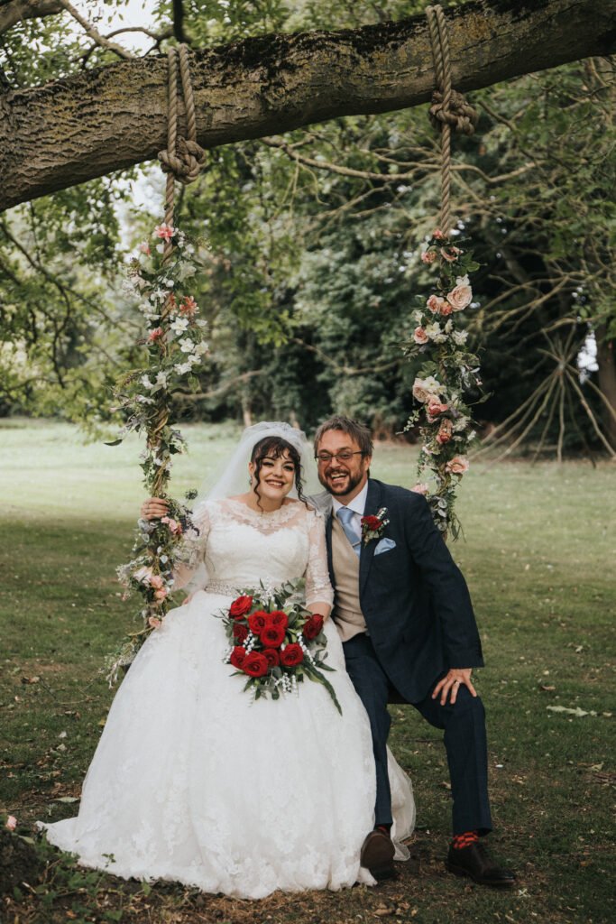 A bride in a white lace gown holding red roses and a groom in a navy suit sit together on a flower-adorned swing hanging from a tree. They are smiling, surrounded by greenery, with trees and grass in the background. © Aimee Lince Photography, Lincolnshire photographer