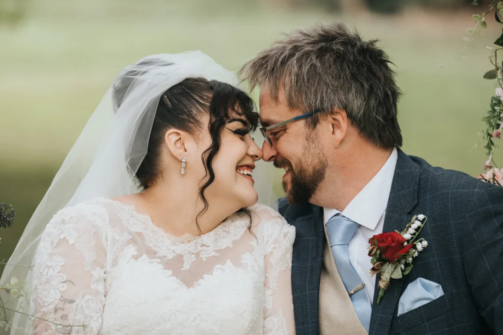 A bride and groom sit closely, smiling and touching noses. The bride wears a lace wedding dress and veil, and the groom wears glasses, a blue suit, and a buttonhole with red and white flowers. They appear joyful and affectionate in an outdoor setting. © Aimee Lince Photography, Lincolnshire photographer