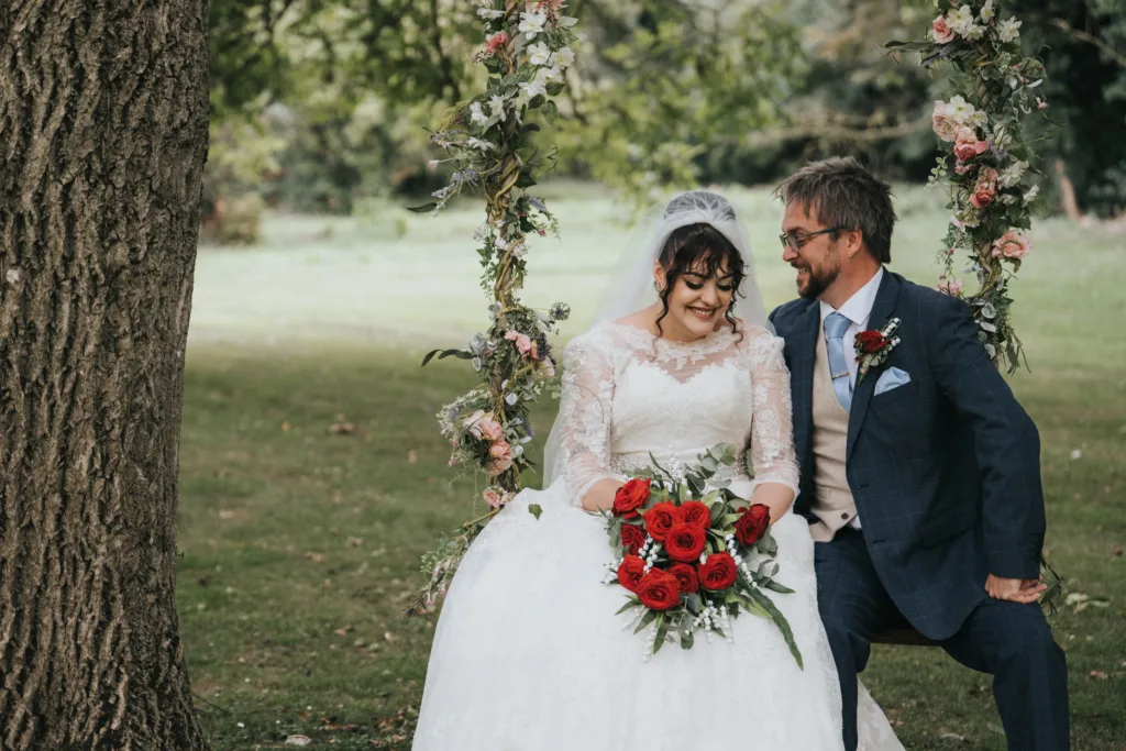 A bride in a white lace gown holding red roses sits on a floral swing next to a groom in a blue suit. They smile at each other in a green outdoor setting with trees and grass. © Aimee Lince Photography, wedding and lifestyle photographer in Lincolnshire